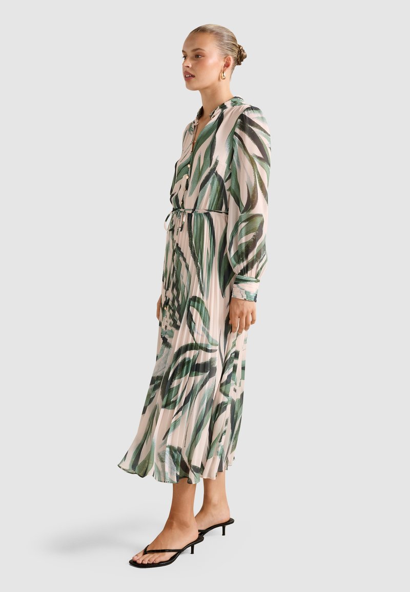 Femme debout de profil portant une robe midi beige avec des motifs de feuilles vertes, des manches longues, un devant boutonné et des sandales à talons noires.