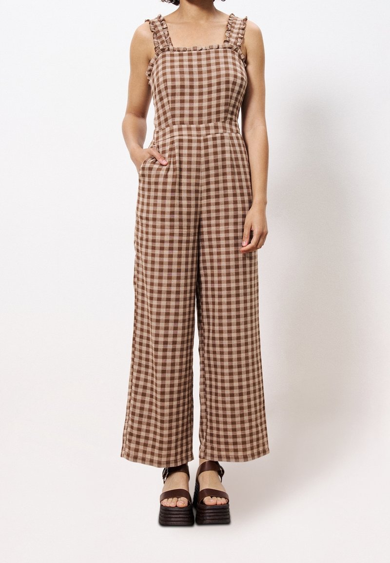Femme portant une combinaison sans manches à carreaux marron et beige avec bretelles à volants et pantalons amples, associée à des sandales compensées.