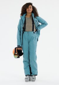 Teal skidjacka med justerbar huva, matchande teal skidbyxor och grå skidstövlar. Modellen håller en orange hjälm med spegelvisir.