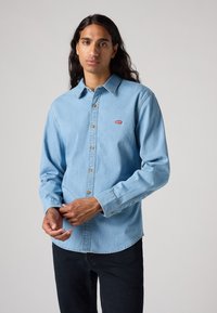 Camicia in denim azzurro chiaro con colletto button-down, maniche lunghe e un piccolo logo rosso di Levi's sul petto. Presenta chiusura a bottoni e orlo curvato.