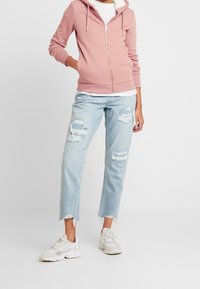 Persona con sudadera de cremallera rosa claro, jeans rasgados azules claros y zapatillas blancas, de pie contra un fondo blanco liso.