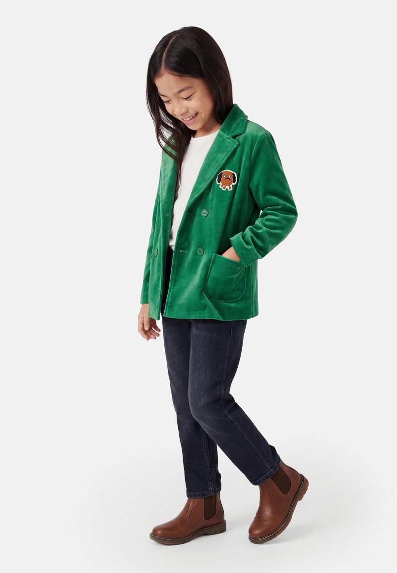 Niña sonriendo, lleva una blazer de terciopelo verde con parche de perro, camisa blanca, vaqueros oscuros y botines marrones, de pie con una mano en el bolsillo.