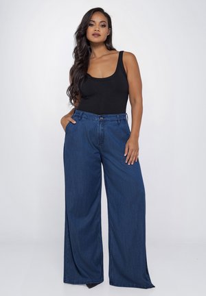 Vrouw met lang donker haar, gekleed in een zwart mouwloos topje en wijde blauwe spijkerbroek, staand met één hand in de zak.