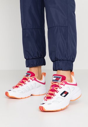White sneakers with orange soles, pink and orange laces, worn with navy blue pants with elastic cuffs.