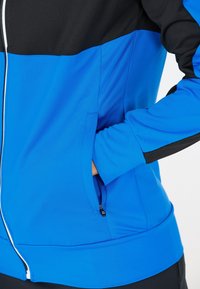 Blå och svart sportig zip-up jacka med en jämn textur, sidofickor, kontrasterande färgblockdesign och åtsittande ärmslut.