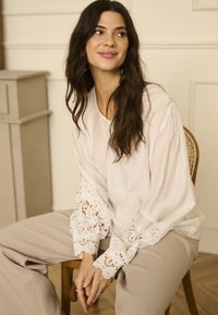 Blusa bianca con maniche rifinite in pizzo, abbinata a pantaloni beige. Il tessuto sembra leggero e texturizzato, con una vestibilità ampia e rilassata.