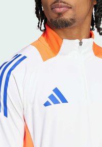 Weiße Sportjacke mit orangefarbenen Akzenten und blauem Streifenmuster. Reißverschluss am Kragen; verfügt über das Adidas-Logo in Blau auf der Brust.