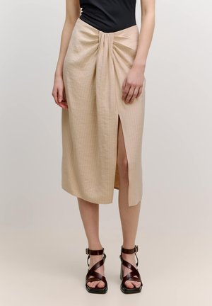 Beige midi skirt with a twisted front detail, featuring a thigh-high slit. Smooth texture and vertical pinstripes. Paired with brown strappy heels.