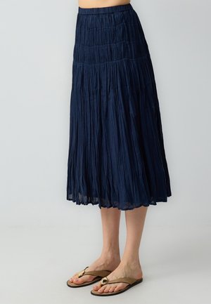 Jupe longue plissée bleu marine avec une taille smockée, fabriquée en tissu léger, présentant une texture fluide et un design longueur cheville.