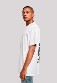 Weißes Baumwoll-T-Shirt mit lässiger Passform, kurzen Ärmeln und großem schwarzen Grafikdruck auf dem Rücken. Das Model wird von der Seite gezeigt.