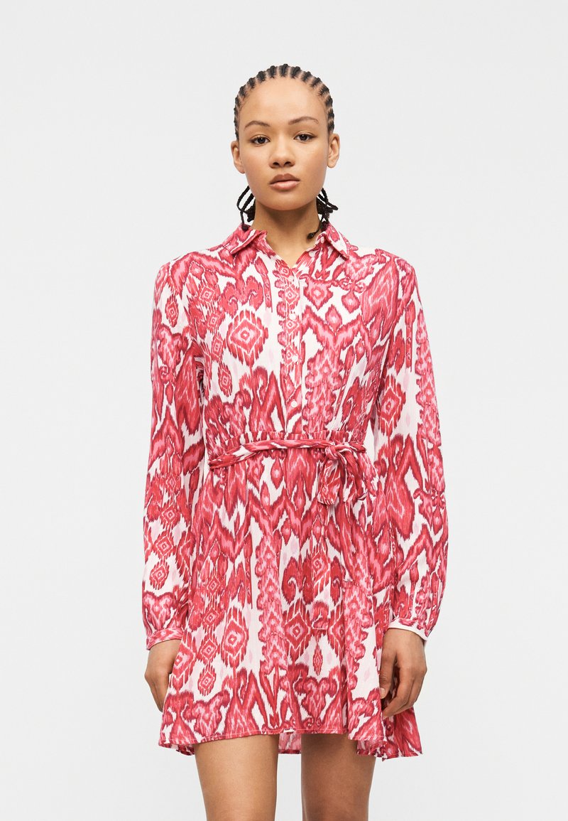 Γυναίκα με πλεγμένα μαλλιά που φοράει κόκκινο και λευκό φόρεμα με σχέδια μέχρι το γόνατο, μακριά μανίκια και ζώνη στη μέση.