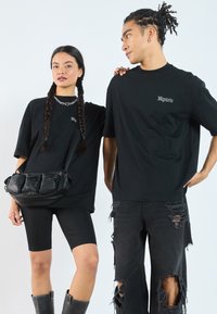 Zwarte, loszittende t-shirts met geborduurde logo's, gecombineerd met zwarte fietsbroekjes en verouderde zwarte jeans. Model draagt een zwarte handtas.