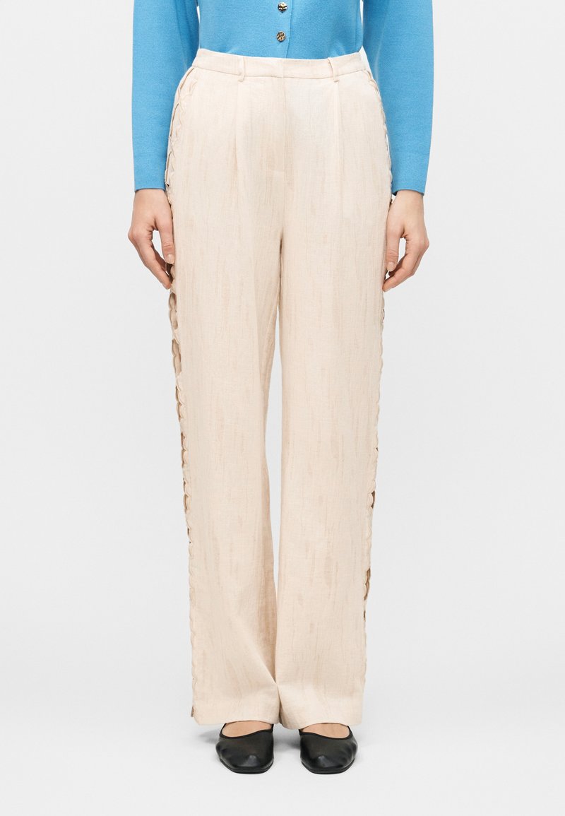 Μπεζ παντελόνια φαρδιάς γραμμής με κυματιστές λεπτομέρειες στα πλαϊνά, φορεμένα με μπλε μπλουζάκι με κουμπιά και μαύρα επίπεδα παπούτσια.