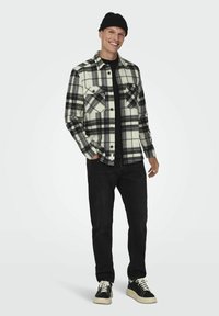 Camisa de franela a cuadros en blanco y negro con dos bolsillos en el pecho, combinada con una camiseta negra, jeans negros y zapatillas negras con suelas blancas.