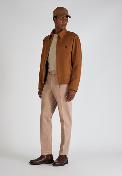 Braune Wolljacke mit einer Vordertasche; beiger Strickpullover; hellrosa Cordhosen; dunkelbraune Stiefeletten; beiger Hut.