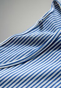 Blå- og hvidstribet stofkrave med en knapdetalje og vaskemærke, der viser en glat tekstur og klare, horisontale linjer.