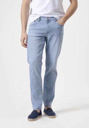 Hombre con vaqueros azul claro, camiseta blanca y zapatos tipo slip-on azul marino, de pie con una mano en el bolsillo.