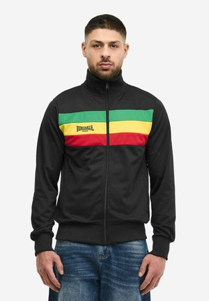 Homme portant une veste noire zippée avec des rayures horizontales vertes, jaunes et rouges et le logo "Lonsdale London", associée à un jean bleu.