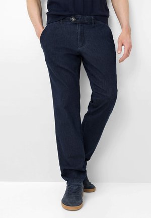 Mann trägt dunkelblaue Jeans mit geradem Schnitt, Hände in den Taschen, kombiniert mit marineblauen Turnschuhen mit hellbraunen Sohlen.