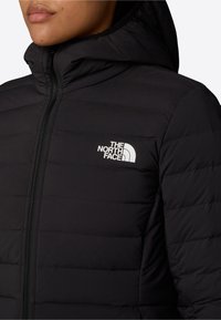 Svart vadderad jacka med huva, med ett förband med dragkedja på framsidan och en vit logotyp på bröstet. Materialet verkar slätt och mjukt.