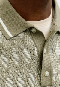 Nahaufnahme einer Person, die ein beiges, gemustertes Strick-Poloshirt mit drei Knöpfen und einem gerippten Kragen über einem weißen T-Shirt mit Rundhalsausschnitt trägt.