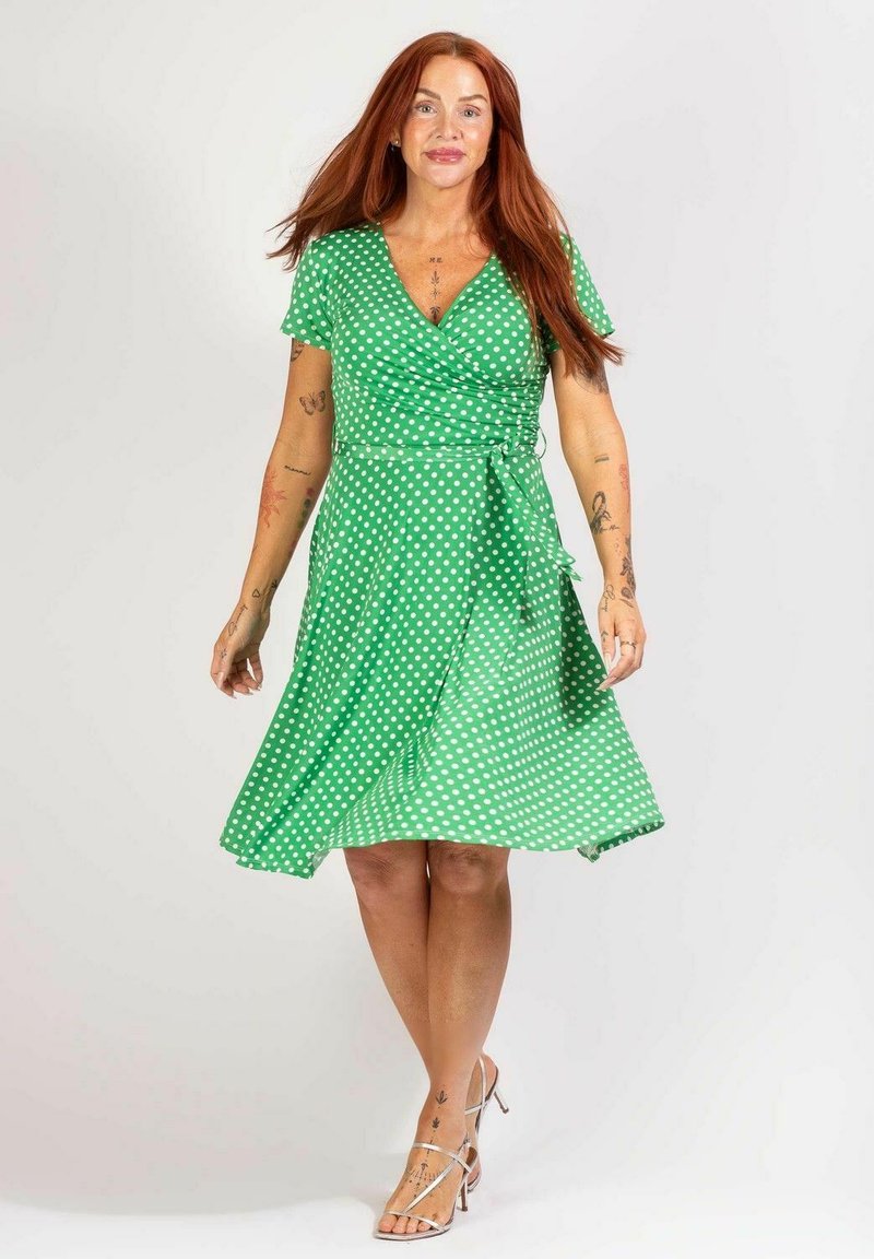 Punatukkainen nainen, joka on pukeutunut vihreään pilkulliseen kietaisumekkoon ja hopeisiin korkokenkiin, seisoo yksinkertaisen valkoisen taustan edessä.