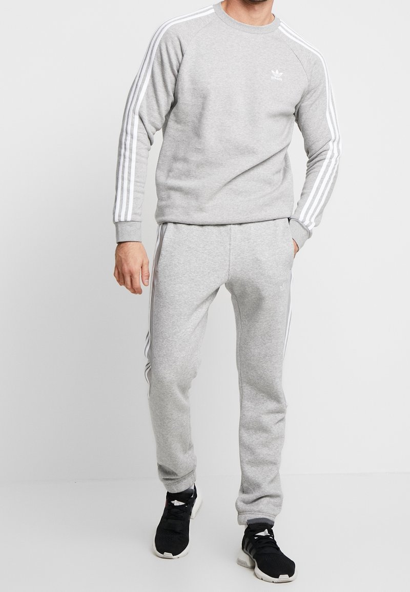 Man som bär en ljusgrå Adidas träningsoverall med vita ränder och svarta Adidas sneakers, står framför en helt vit bakgrund.