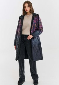 Žena oblečená v dlouhém prošívaném kabátu s fialovými květinovými vzory, béžovém svetru, černých džínách a černých botách, stojící na jednobarevném pozadí.