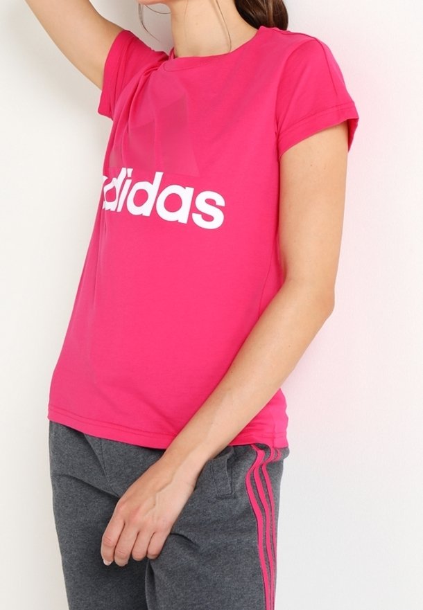 Różowa bawełniana koszulka z podwiniętymi rękawami i białym logo "Adidas" na klatce piersiowej. Noszona z szarymi spodniami dresowymi z różowymi paskami.