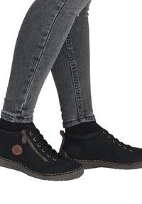 Bottes de cheville en cuir noir avec un bout rond, semelle texturée, fermeture éclair dorée et détails de couture, portées avec un jean skinny gris.