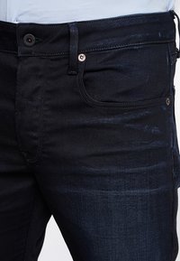 Primer plano de una persona que lleva jeans de mezclilla azul oscuro con botones visibles, presillas para cinturón y detalles del bolsillo delantero.