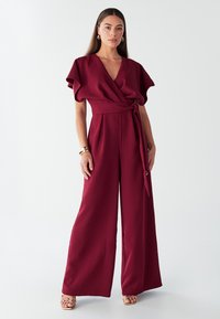 Bordo kombinezon širokih hlačnic z ovitnim dizajnom, v-izrezom in kratkimi krilci na rokavih. Pas je poudarjen z vrvico, tkanina pa je gladka na otip.