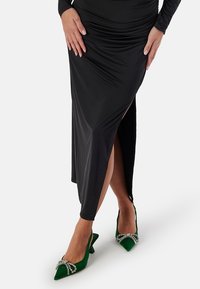Svart, åtsittande klänning med en hög slits till låret, parat med gröna sammetsklackar med en juvelaccent. Slät textur och elegant design.