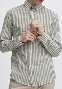 Camisa de manga larga con rayas verticales alternas en verde y blanco, con puños abotonados y cuello. La tela parece tener textura.