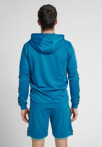 Turkis atletisk hættetrøje med et tekstureret stof, der har sorte diagonale striber på ærmerne. Matcher med shorts i samme farve.