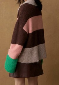 Femme portant un pull en peluche coloré avec des sections marron, rose, gris et vert, associé à une jupe marron foncé sur un fond uni.