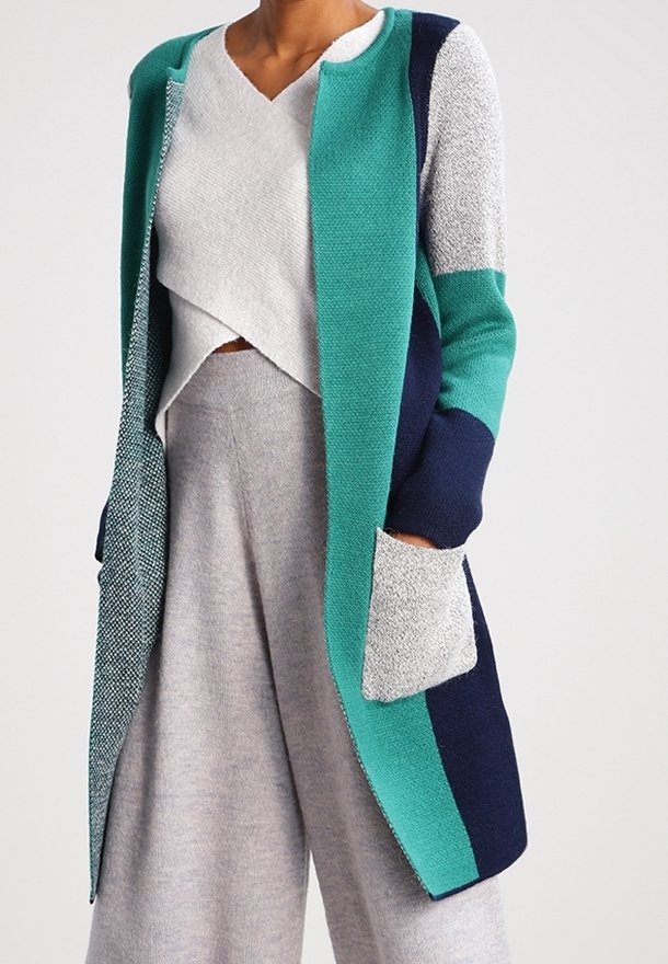 Femme portant un long cardigan color block avec des sections turquoise, marine et gris sur un pull col V gris clair et un pantalon à jambes larges.
