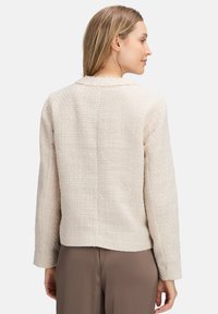 Veste textur�ée beige clair au design court, avec des manches longues et un col rond. Comprend un motif en tissu tissé et des détails de couture subtils.