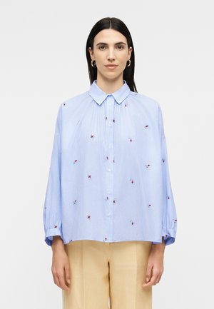 Femme aux cheveux lisses et foncés, portant une chemise à rayures bleues et blanches avec de petits motifs floraux rouges et un pantalon beige, fond neutre.