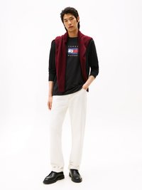 Černá dlouhá halenka s grafickým logem, na které je navlečený vínově červený bezrukávový svetr. Bílé kalhoty a černé boty doplňují celý outfit.