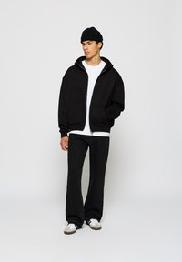 Haut de survêtement zippé noir avec une poche avant, porté sur un T-shirt blanc, associé à un pantalon large noir et des baskets blanches, complété par un bonnet noir.