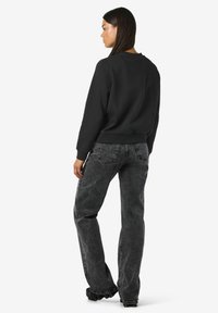 Mujer con pelo oscuro y liso, vistiendo un sudadera negra y jeans negros desgastados, de pie y de espaldas contra un fondo blanco.