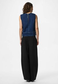 Denim ärmlös topp med dragkedja på ryggen, kombinerad med höga svarta vida byxor. Slät textur, djup blå färg och strukturerad passform.