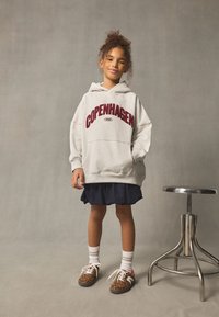 Grauer übergroßer Kapuzenpullover mit der Aufschrift "COPENHAGEN" in kastanienroter Schrift, kombiniert mit einem marineblauen Rock. Braune Schuhe mit Leopardenmuster und weiße Socken vervollständigen das Outfit.