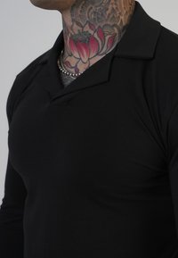 Crna dugosleeve polo majica s klasičnim ovratnikom i glatkom teksturom. Suptilan dizajn, uglavnom jednobojan bez vidljivih uzoraka ili akcenta.
