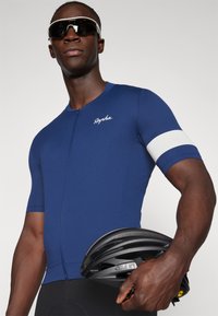 Jersey de ciclismo navy con una superficie texturizada, detalle blanco en la manga, mangas cortas y un cierre frontal. Casco de bicicleta negro sujetado con la mano.