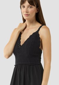 Schwarzes Spitzenkleid mit einem V-Ausschnitt und gewelltem Saum, schmalen Trägern und einem taillierten Oberteil, das in einen fließenden Rock übergeht.