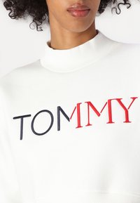Fehér pulóver magas, bordázott nyakkal. Piros és sötétkék hímzett "TOMMY" felirattal rendelkezik, amely modern, laza dizájnt mutat. 