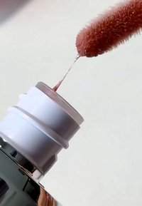 Glänzende braune Flüssigkeit auf einer roten, weichen Applikatorspitze, die in eine Flasche mit weißer Kappe eingetaucht wird, vor einem schlichten Hintergrund.