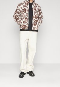 Bílá fleecová bunda s hnědým paisley vzorem, zapínání na zip, nošená přes černé tričko, v kombinaci s bílými kalhotami a černými botami.